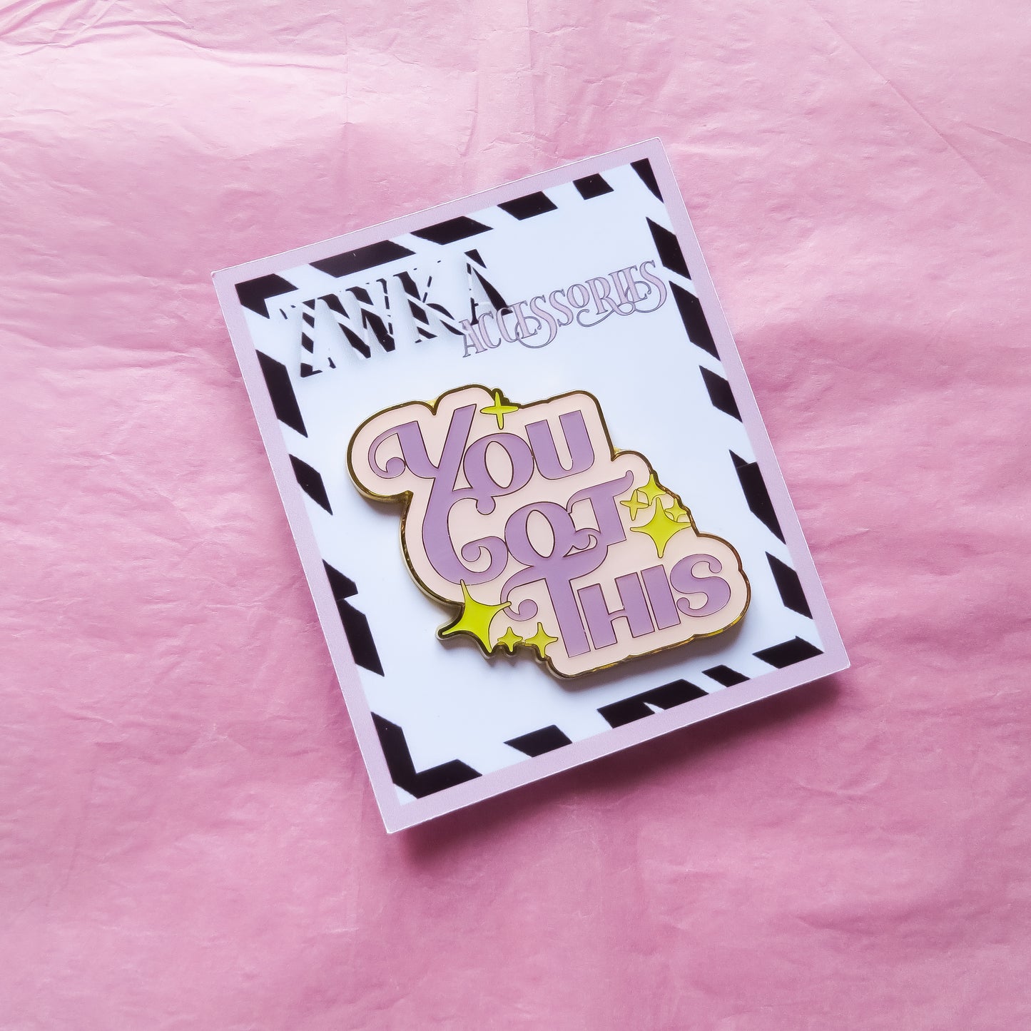 A pastel enamel pin with the words "You Got This" in bold lettering on a backing card on pink tissue paper.