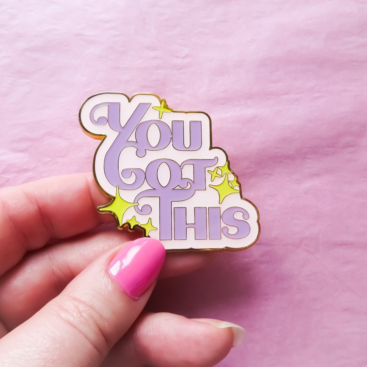A pink and purple enamel pin with the words "You Got This" in bold lettering being held with pink tissue paper as a background.