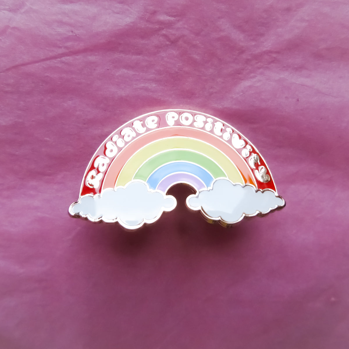 Rainbow enamel pin featuring a rainbow and two clouds with the words ‘Radiate Positivity’ in gold-plated hard enamel, 2 inches wide.