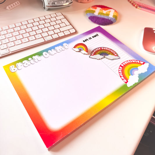 Colourful rainbow brain dump A4 notepad with rainbow punch needle mug rug and enamel pin and matching glitter sticker on a desk with keyboard and mouse.