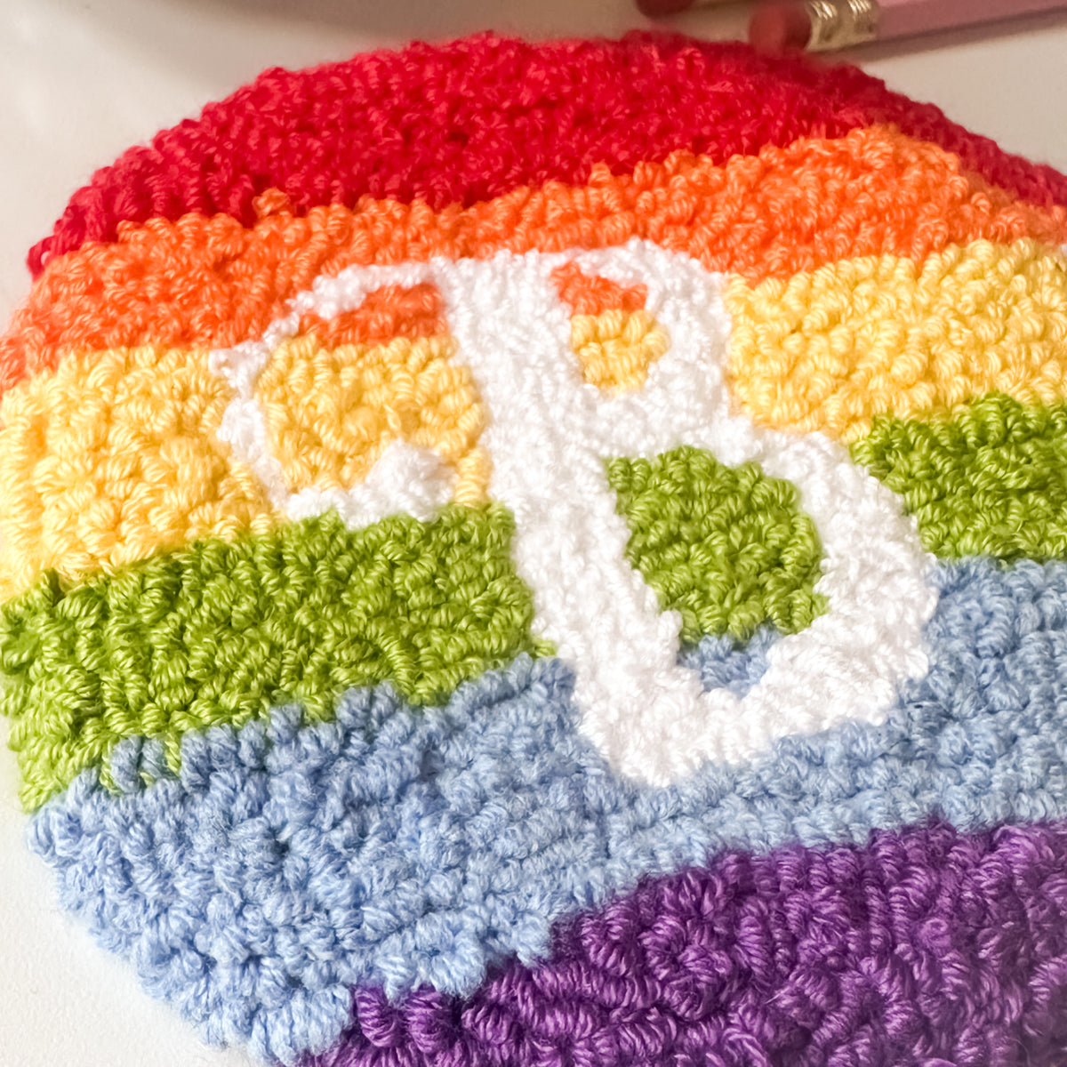Detailed close up view of rainbow mug rug yarn texture – round punch needle coaster in bright rainbow colours with the letter B in white yarn.