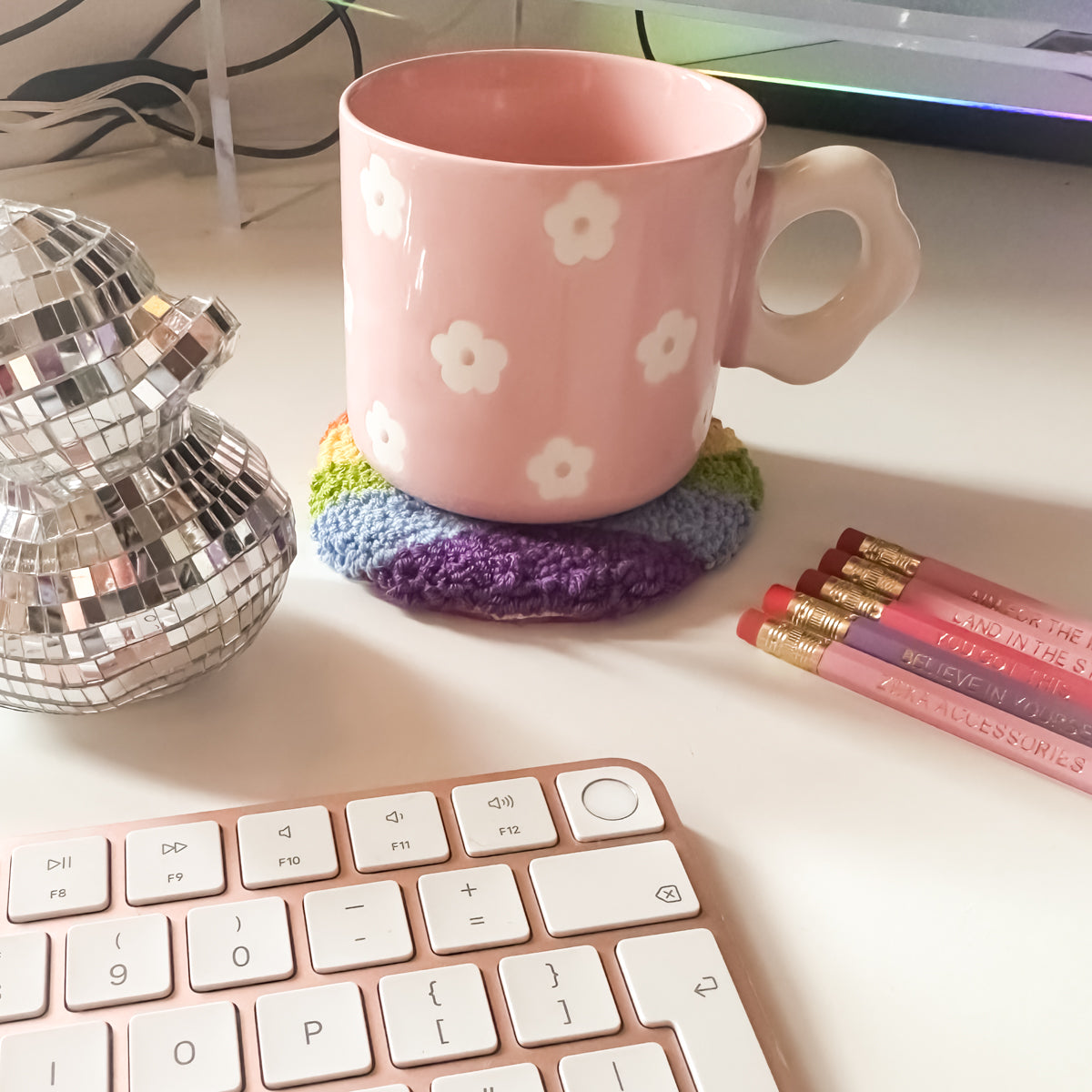 Colourful rainbow punch needle mug rug with pink ceramic mug on top – cosy handmade coaster for hot drinks.