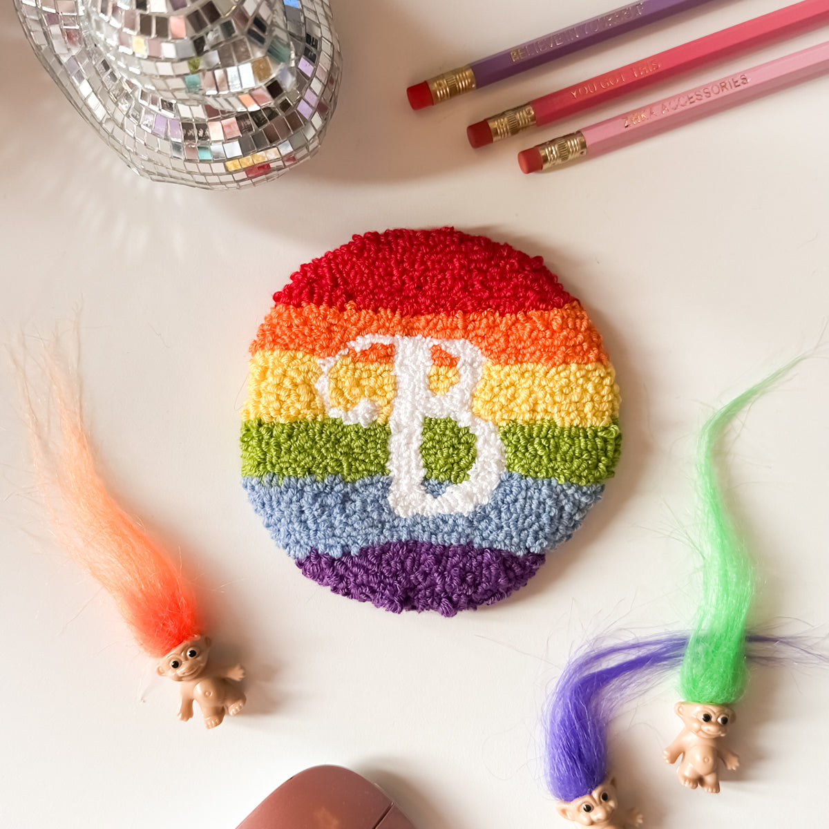 Handmade rainbow round mug rug coaster in bright colours with letter B in white on a white desk surface – 50% acrylic 50% cotton blend, approx. 11cm.