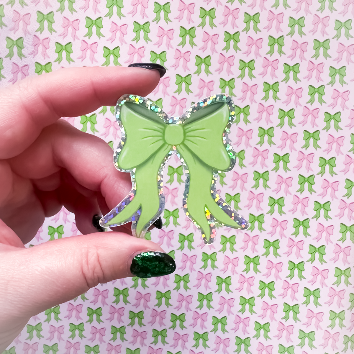 Hand holding a green bow-shaped glitter sticker against a patterned background with green and pink bows.