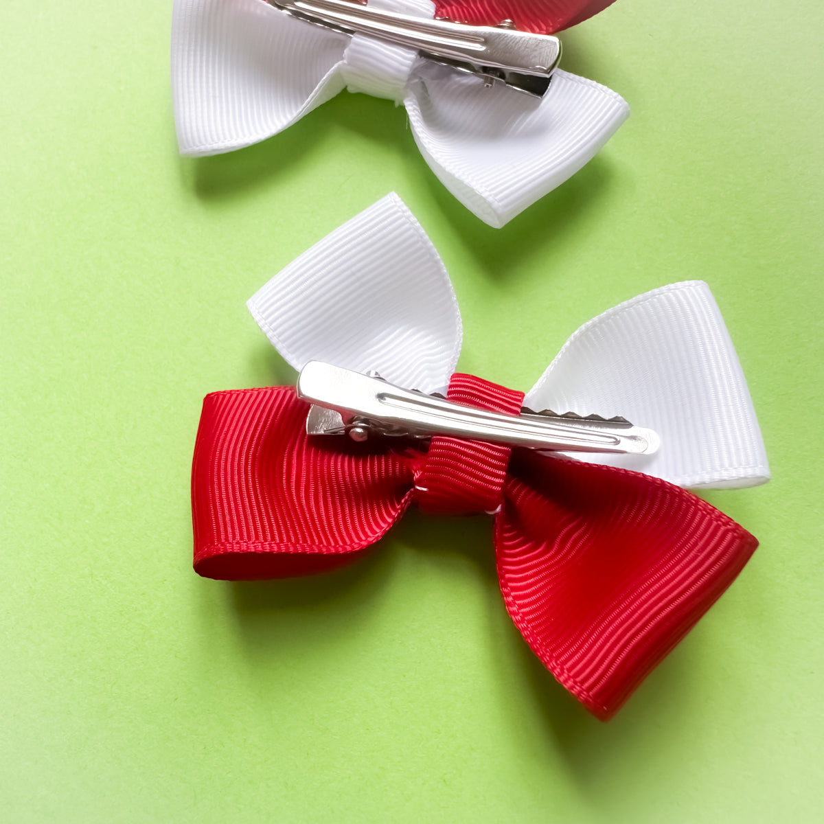 Two hair bows, one red and one white, with metal alligator clips on a green background.