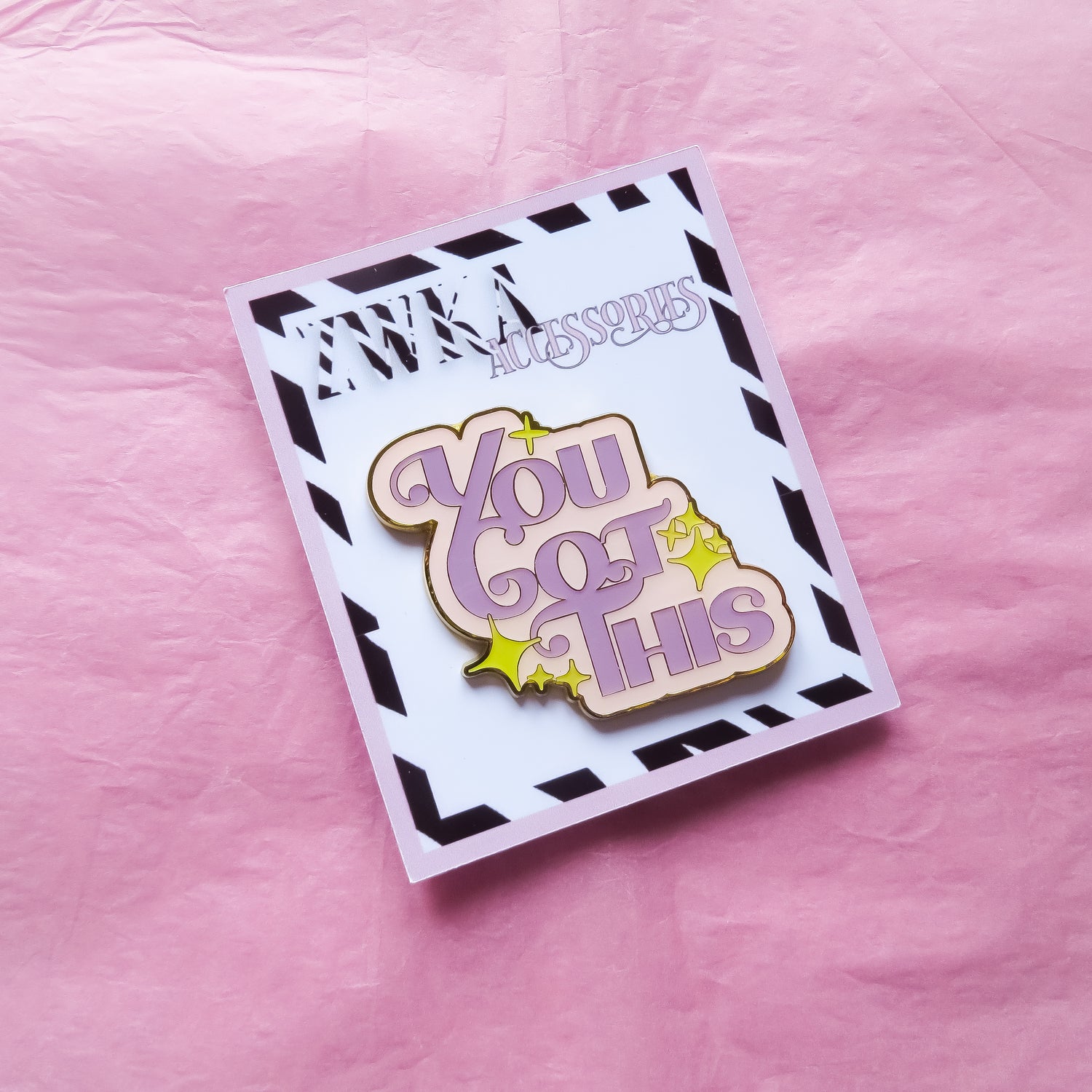 A pastel enamel pin with the words "You Got This" in bold lettering on a backing card on pink tissue paper.