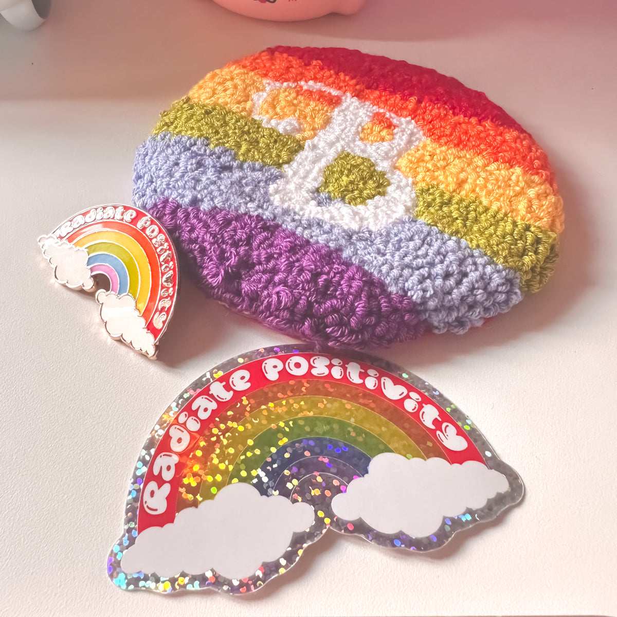 Colourful punch needle round mug rug with rainbow stripes and 'Radiate Positivity' Rainbow shaped enamel pin with matching glitter sticker on a white background.
