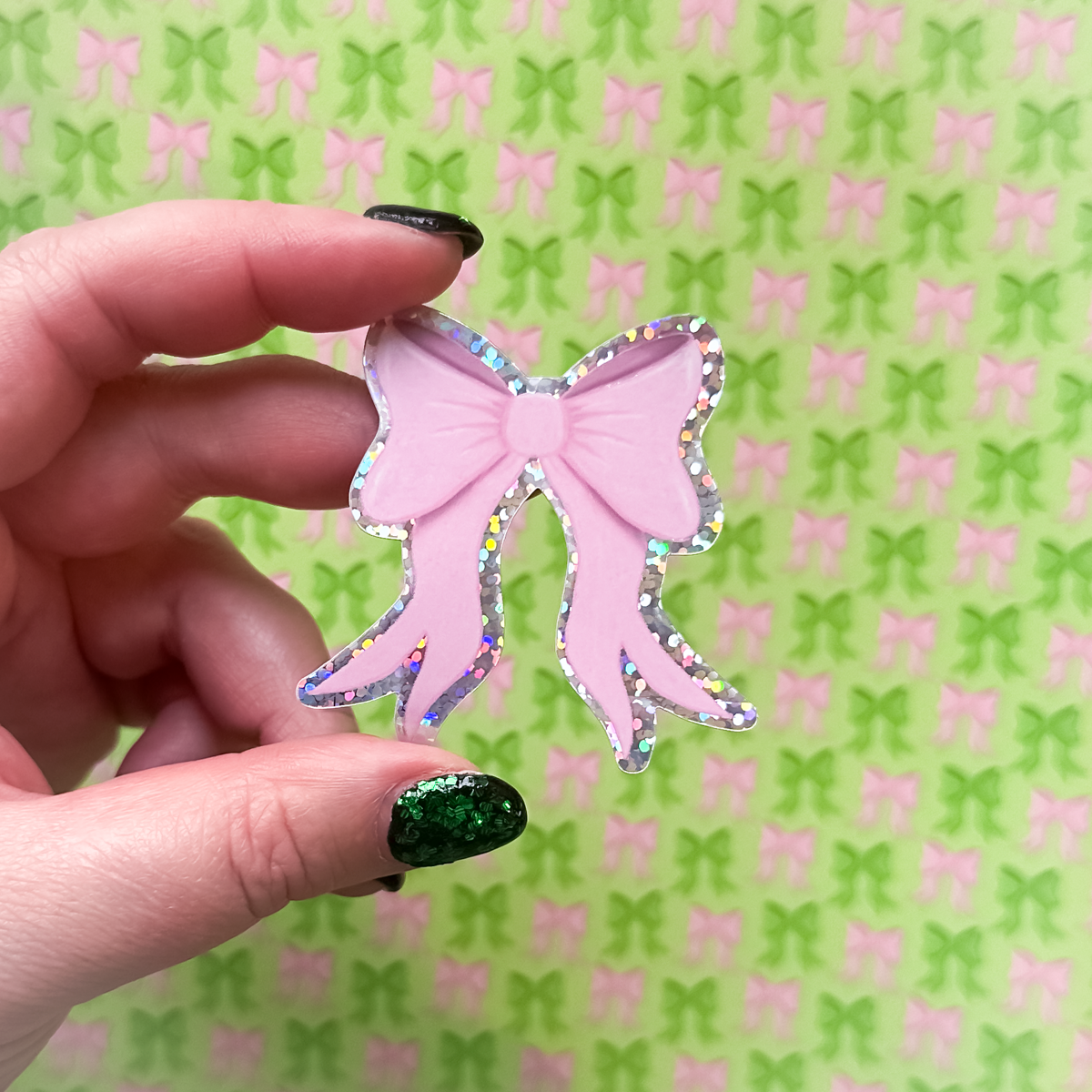 Hand holding a pink bow-shaped sticker with iridescent glitter against a patterned background with pink and green bows.