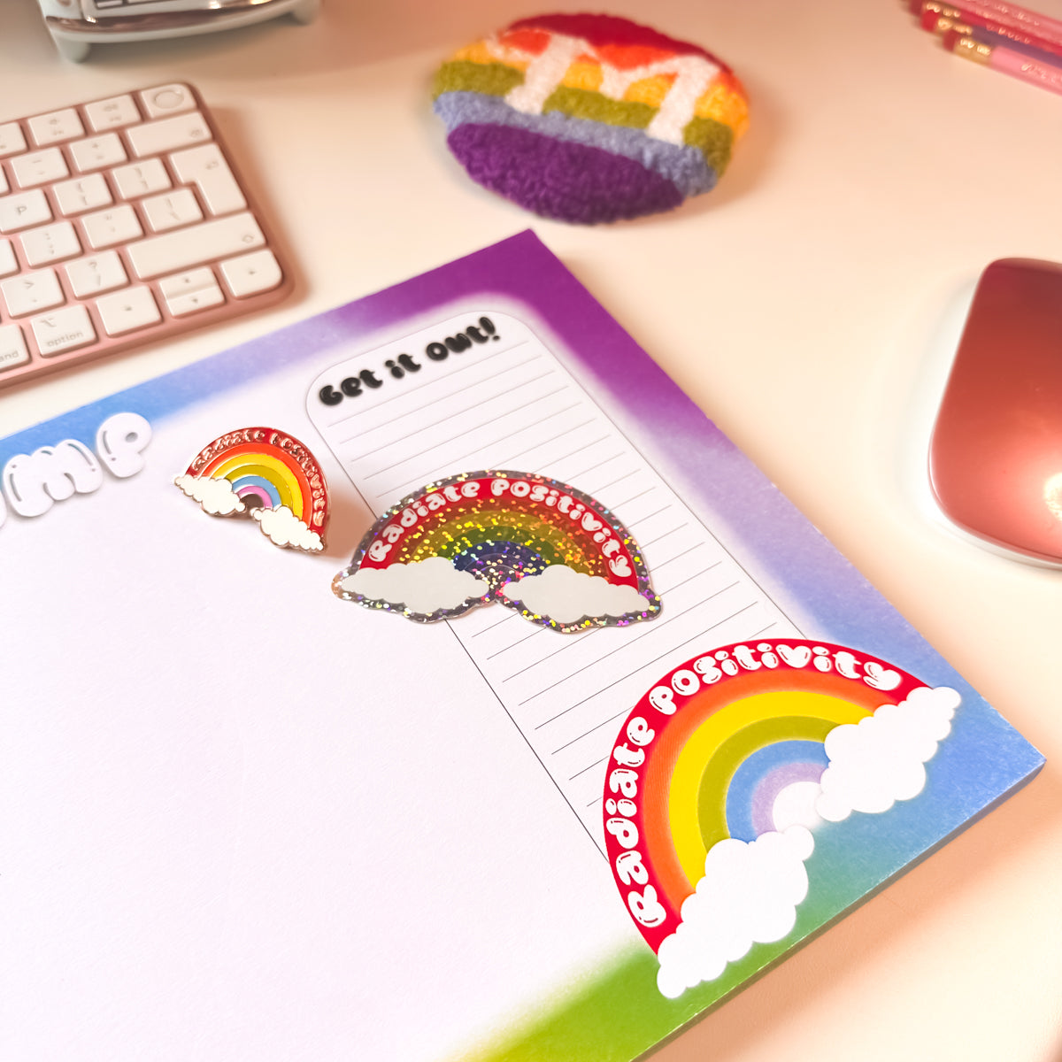 Colourful rainbow-themed items including a punch needle rainbow initial mug rug, a rainbow enamel pin, and a matching glitter sticker on a 'Brain Dump' notebook on a desk with keyboard and mouse.