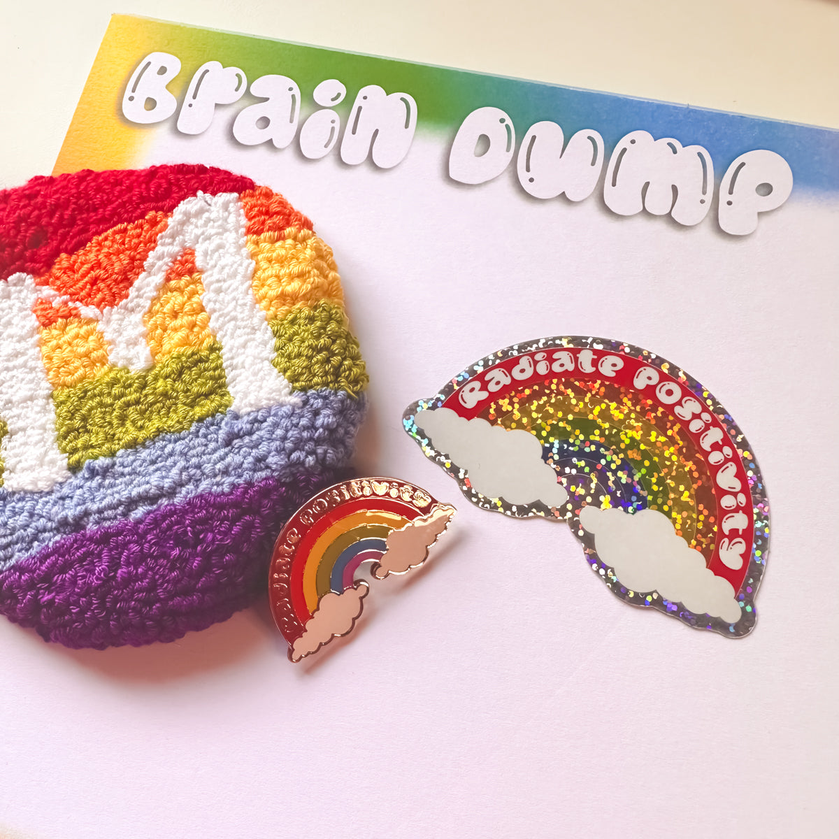 Colourful rainbow-themed items including a punch needle rainbow initial mug rug, a rainbow enamel pin, and a matching glitter sticker on a 'Brain Dump' notebook.