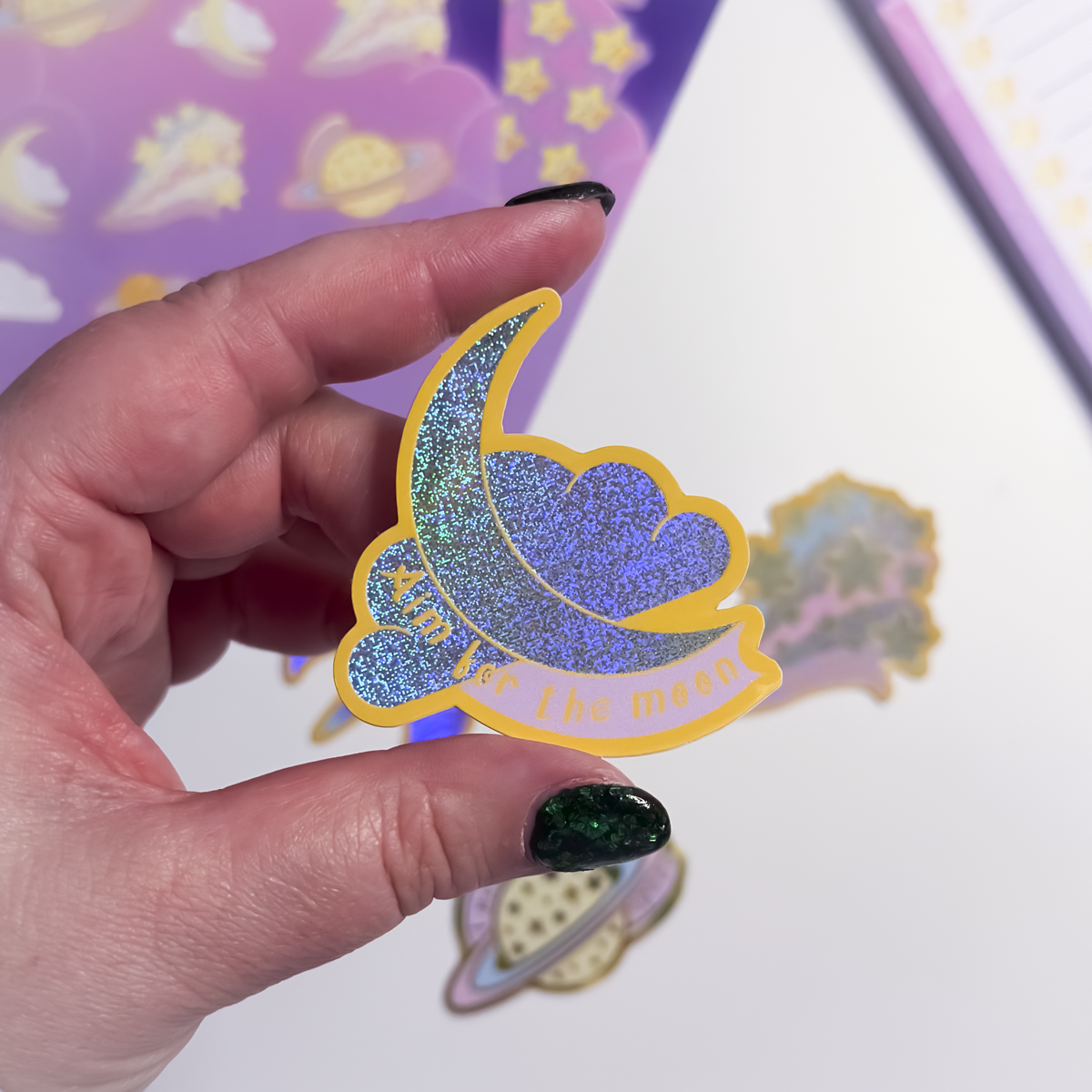 Hand holding a glittery sticker with a moon and star design against a colorful background