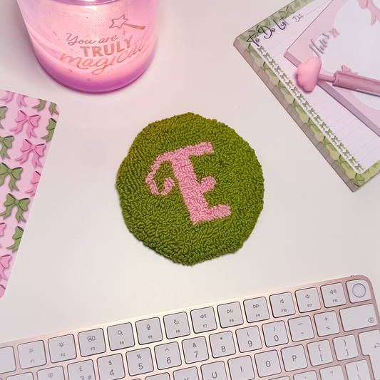 Green textured mug rug coaster with pink letter 'E' on a white surface with a keyboard and decorative items.