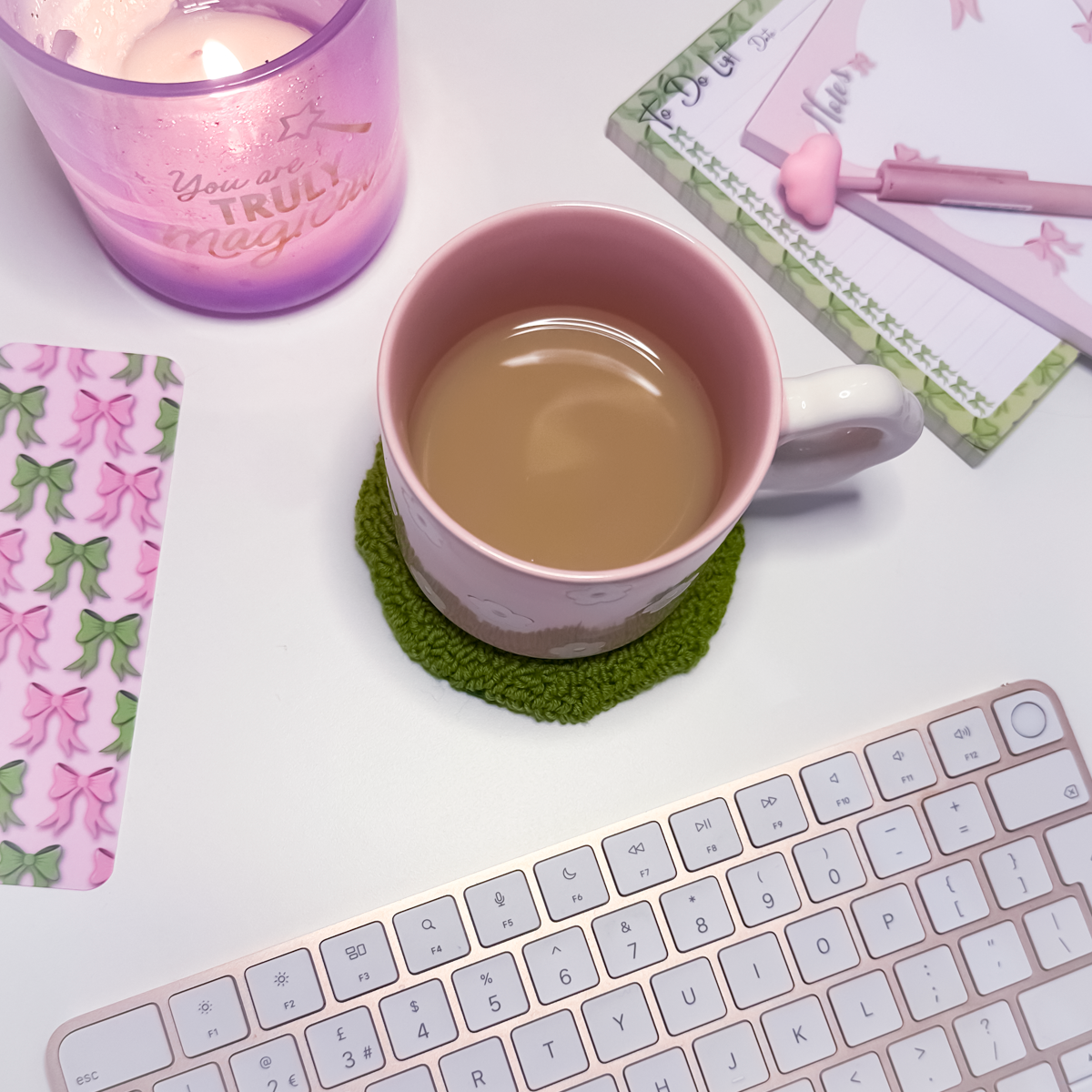 Pink mug with tea on a green coaster next to a keyboard, candle, and stationery items on a white surface.