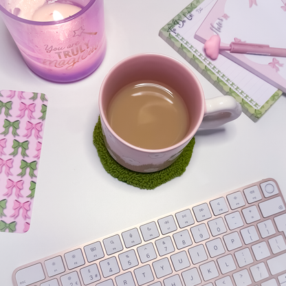 Pink mug with tea on a green coaster next to a keyboard, candle, and stationery items on a white surface.