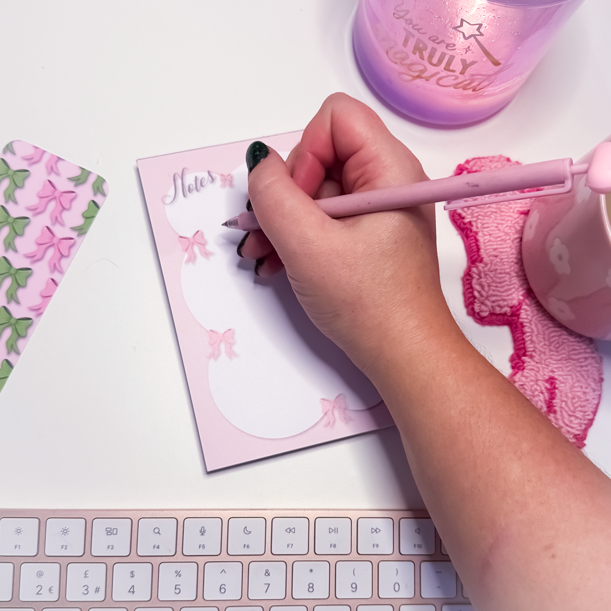 Hand writing on a pink notepad with a pink pen, surrounded by stationery items.