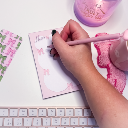 Hand writing on a pink notepad with a pink pen, surrounded by stationery items.