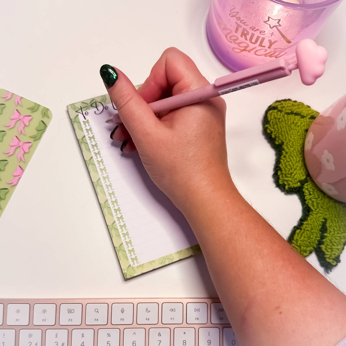 Hand writing on a green bows A6 notepad with a pink pen, surrounded by stationery items on a desk.