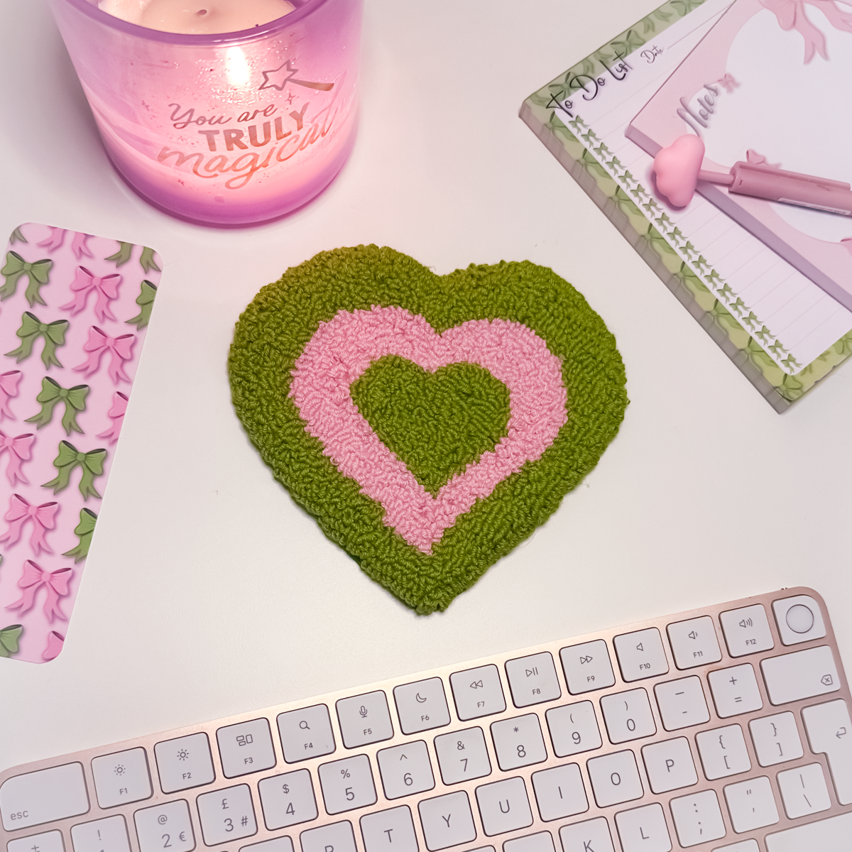 Heart-shaped green and pink mug rug on a desk with a candle and stationery items.