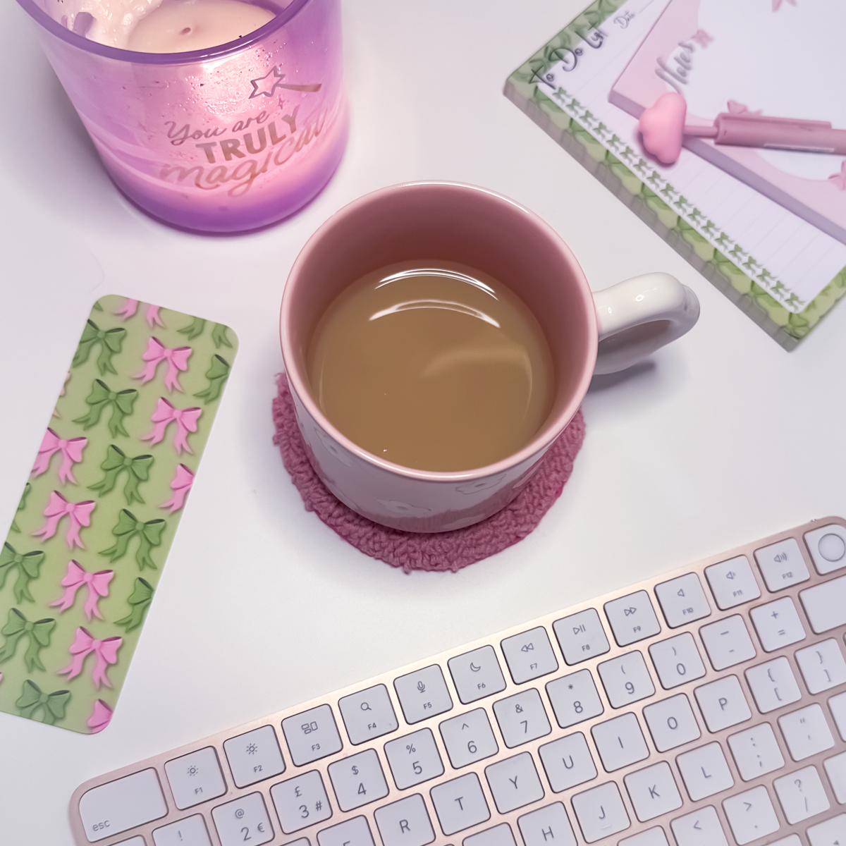 Pink mug with tea on a pink round mug rug on a white surface with a keyboard and decorative items.