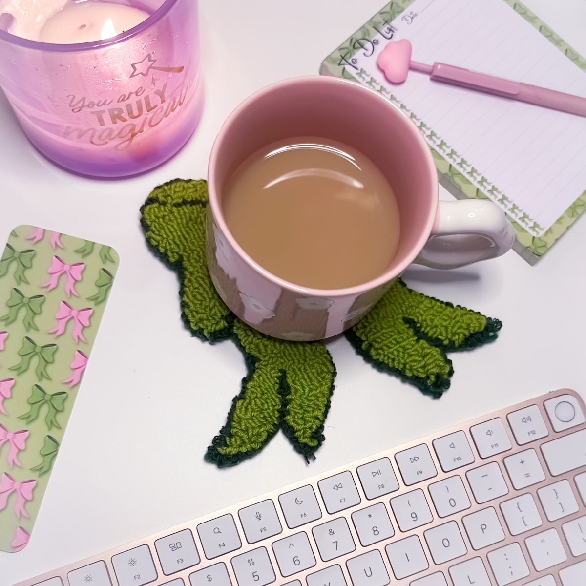 Pink mug with a bow-shaped mug rug coaster on a desk with a keyboard and stationery items.