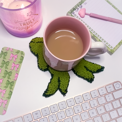Pink mug with a bow-shaped mug rug coaster on a desk with a keyboard and stationery items.