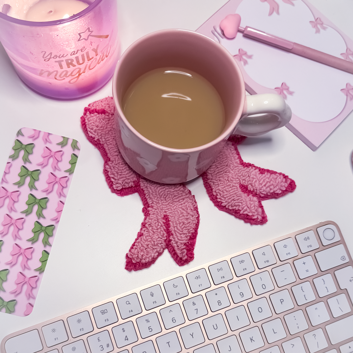 Pink mug with tea on a decorative pink bow mug rug coaster next to a keyboard, candle, and stationery items.