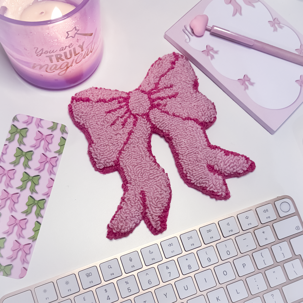 Pink bow mug rug on a white surface with a candle, notebook, and keyboard.